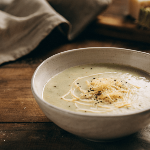 Bowl of creamy courgette (zucchini), potato and cheddar soup topped with grated cheese and black pepper on a rustic wooden table.